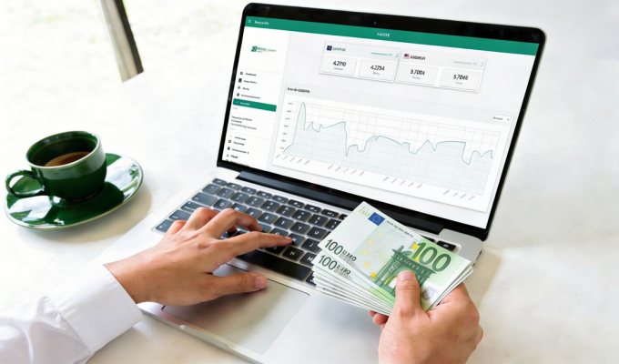 Laptop z kursem walut na ekranie, plik banknotów 100 euro w prawej dłoni, zielona filiżanka z kawą